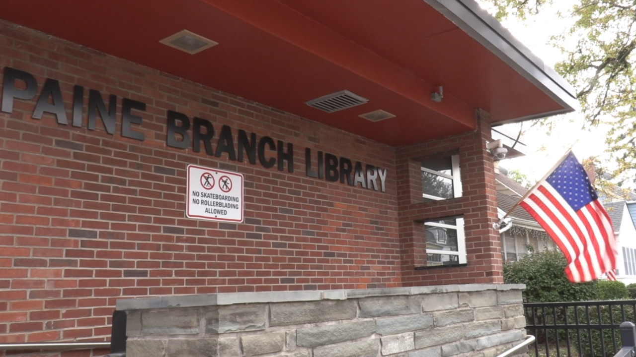 The brick font wall of the paine branch library. In big black letters it says "Paine Branch Library." On the right of the photo the us flag waves. It's a cloudy overcast day.